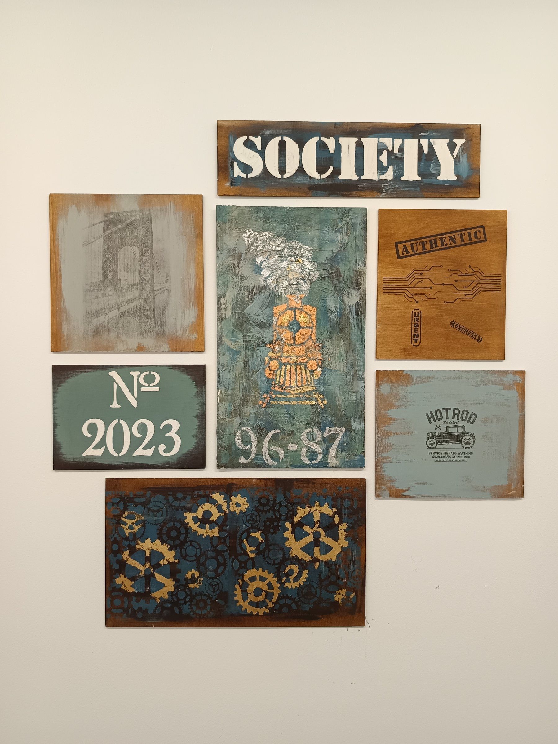 Composizione di tavole dipinte su parete bianca: scritte a stencil, ingranaggi dorati, texture verdi e legno